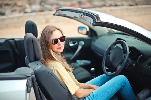 A teenage girl sits behind the wheel of her first car. The top is down and she is wearing sunglasses.